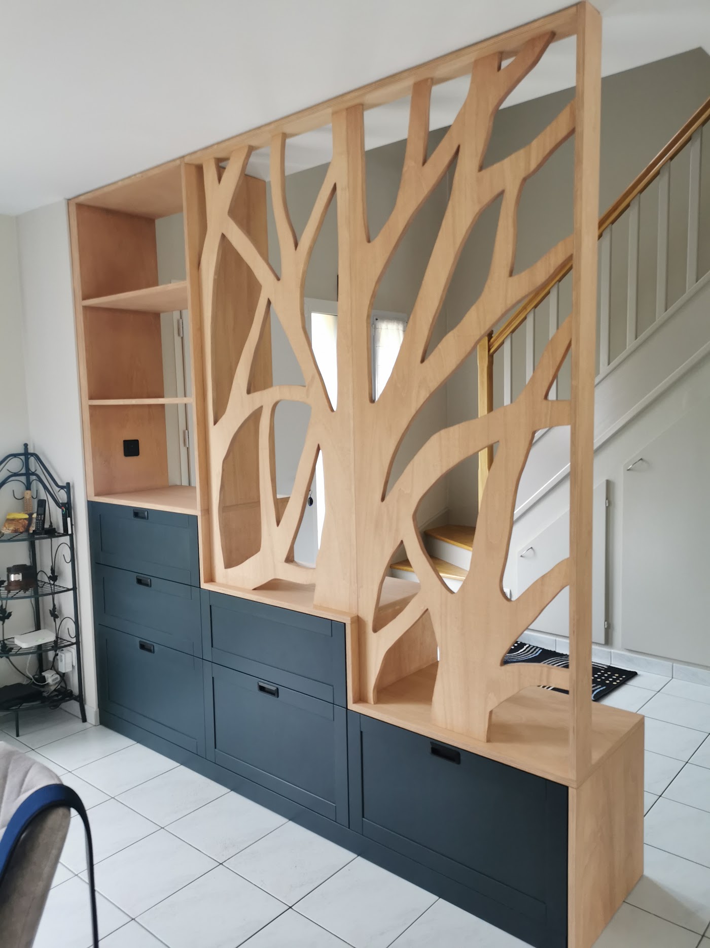 Claustra en bois découpé en forme d'arbre, posé sur un meuble à tiroirs, sous un escalier.